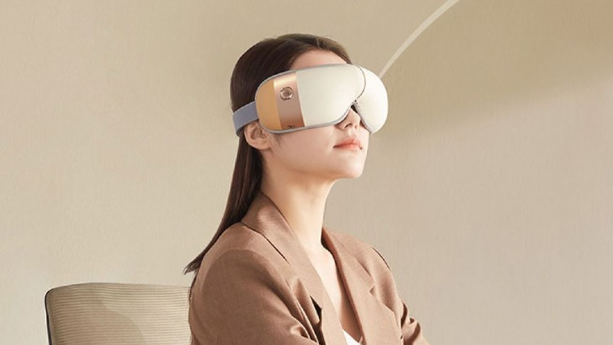 Người ngồi trên ghế, đeo thiết bị massage mắt hiện đại, nền màu be nhạt.